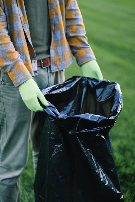 A person standing outdoors on a grassy area is shown holding a large, partially open black rubbish bag made of plastic, with a glossy finish reflecting natural light. The individual is wearing a yello
